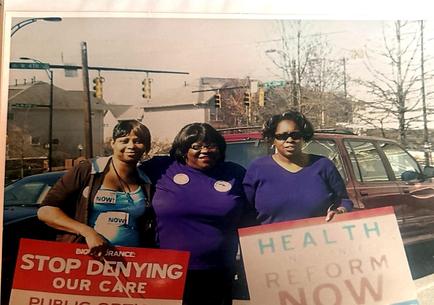 Three community advocates on a Charlotte street holding signs reading 'Stop Denying Our Care' and 'Health Reform Now'.