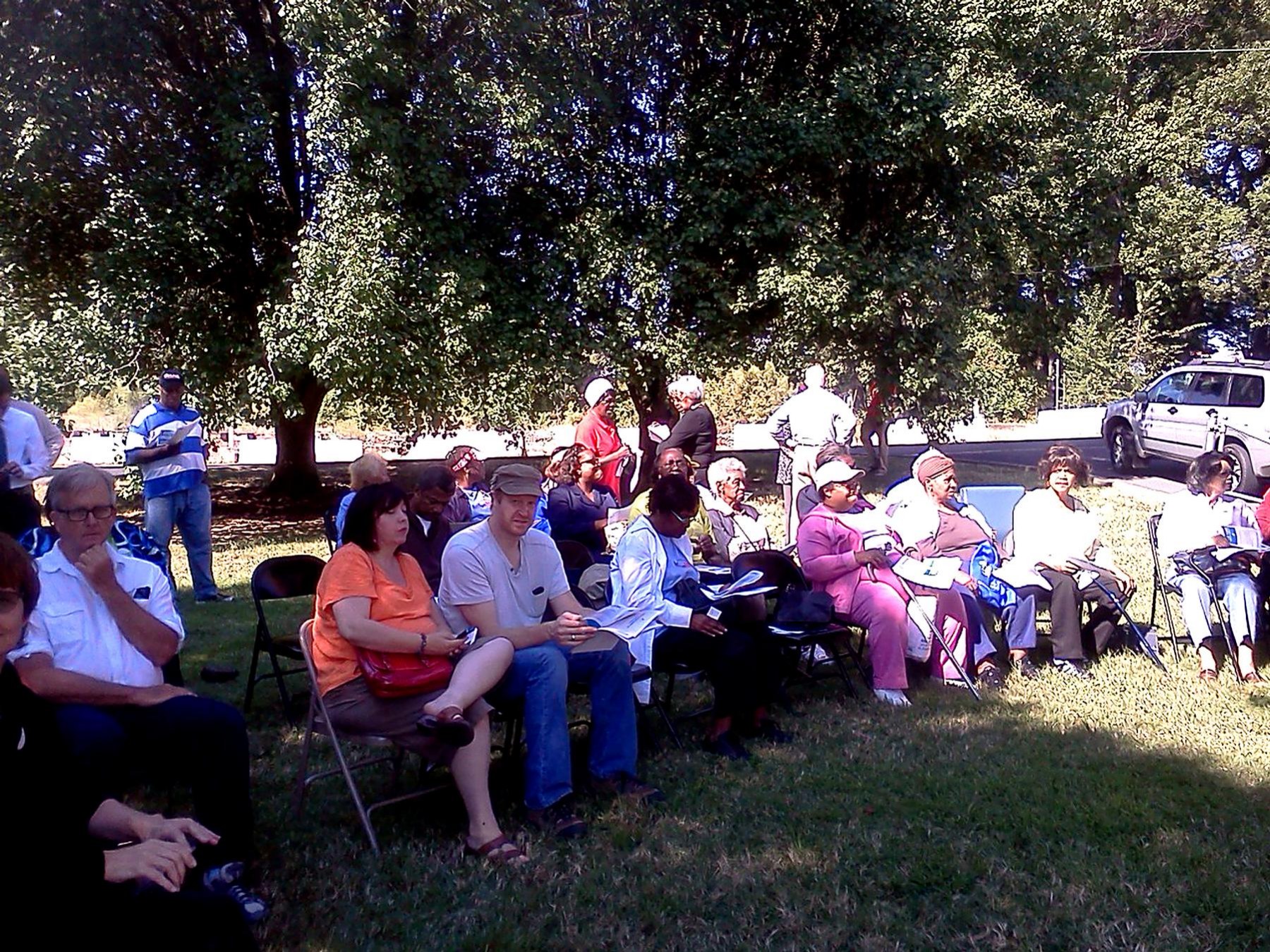 Community members seated in folding chairs under shade trees listening at an outdoor healthcare gathering.