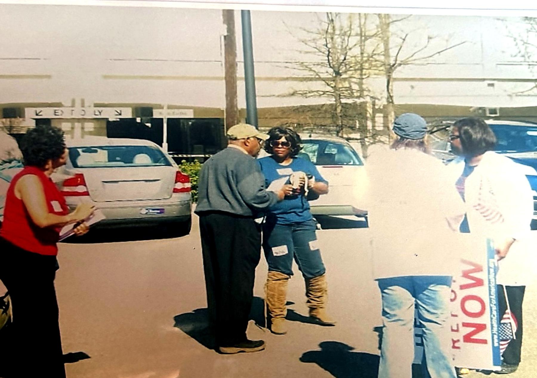 Community members talking beside parked cars while holding healthcare reform signs.