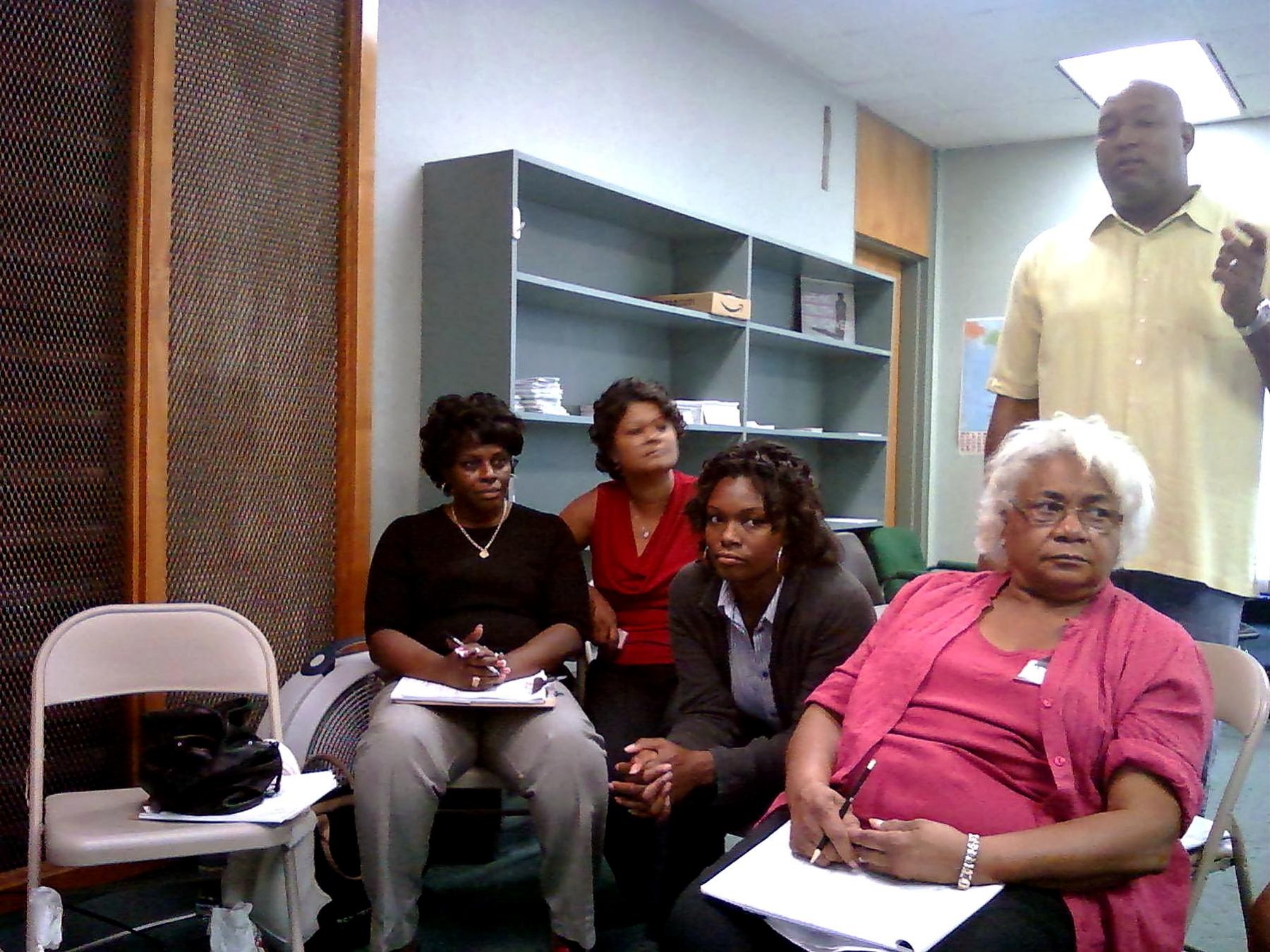 Community members seated with papers in a meeting room while a facilitator stands nearby.
