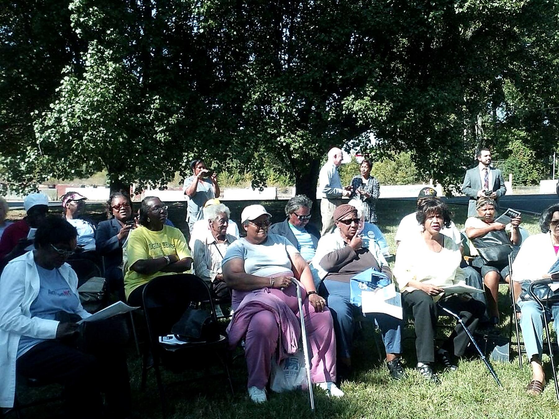 A large group of community members seated outdoors in the shade during a neighborhood healthcare meeting.
