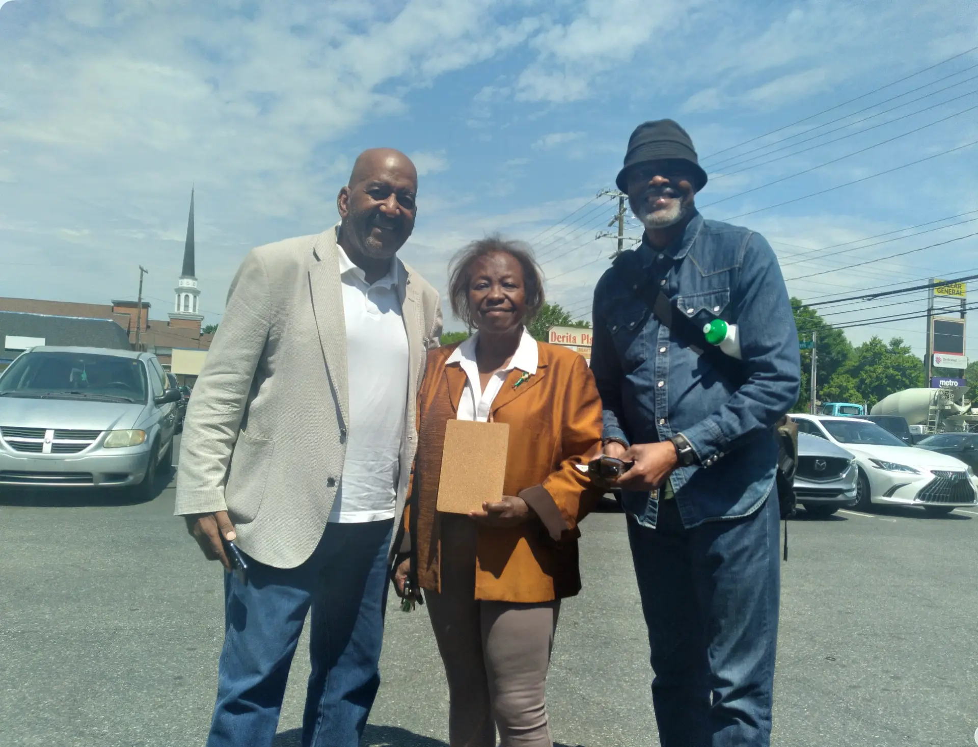Three CHCC community members standing together in a Charlotte shopping-center parking lot on a sunny day, one holding a clipboard, with a church steeple in the background.