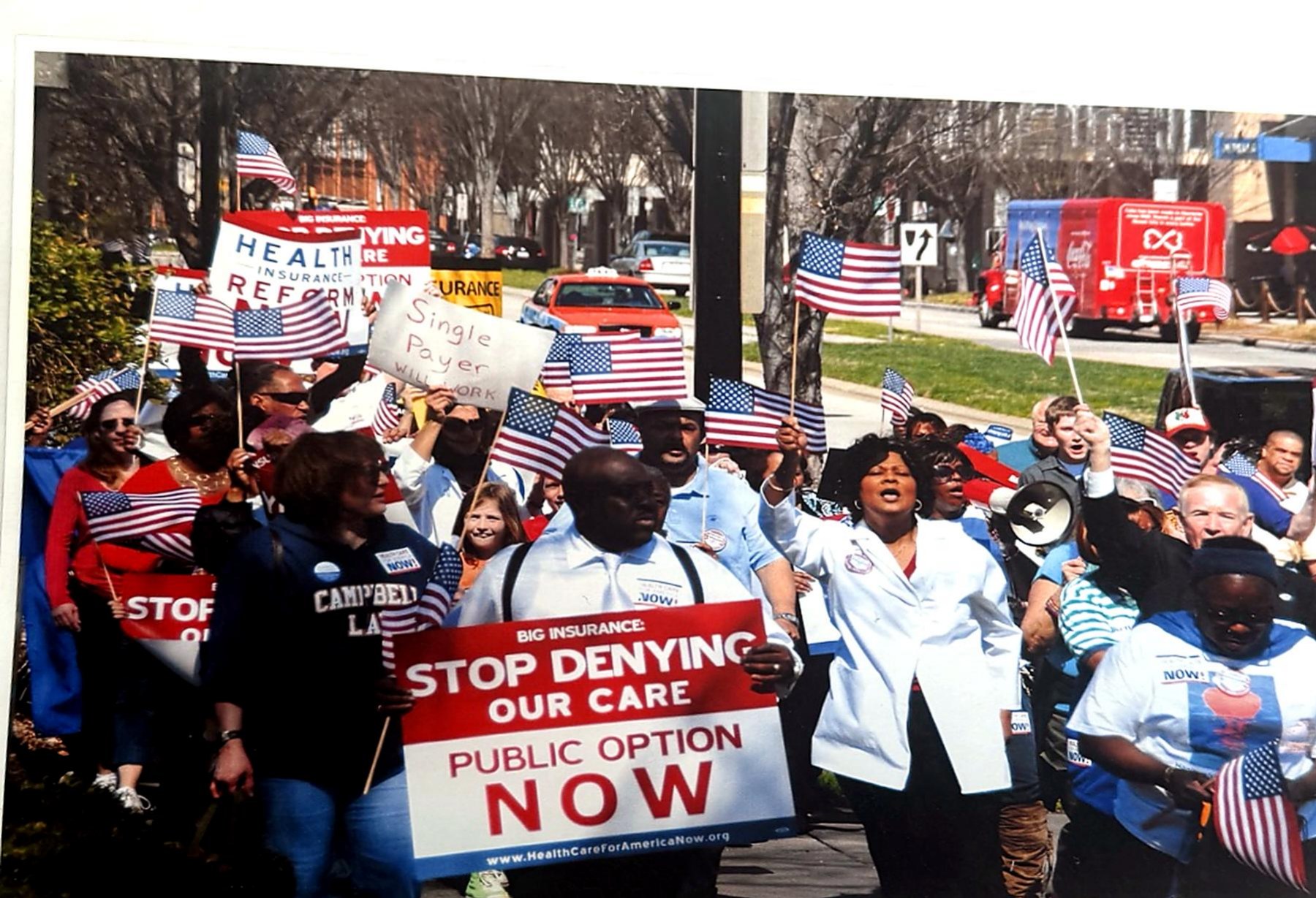 A large crowd of demonstrators holding American flags and 'Public Option Now' signs at a healthcare rally.