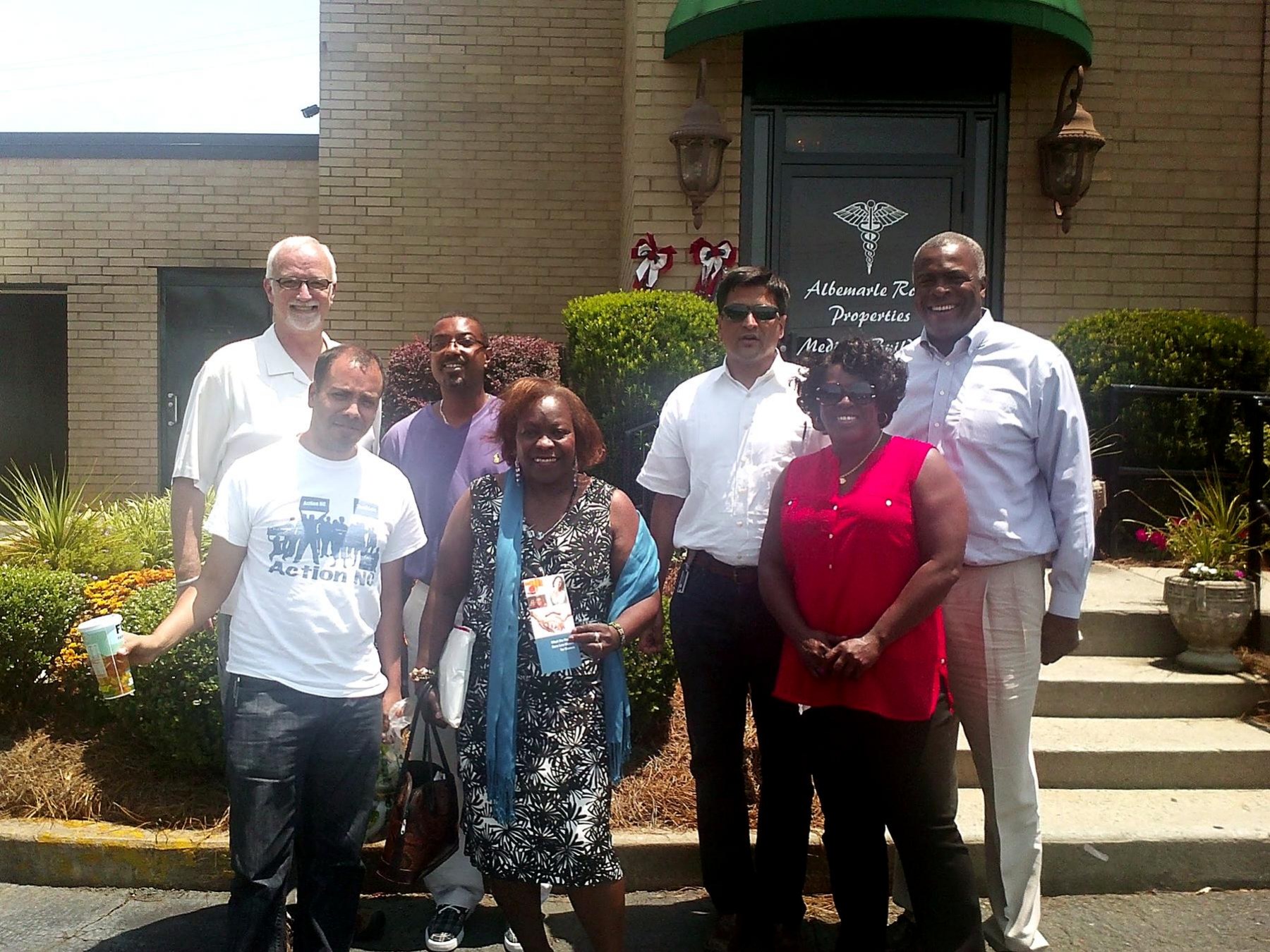 A group of community members standing together outside a brick office building on a sunny day.