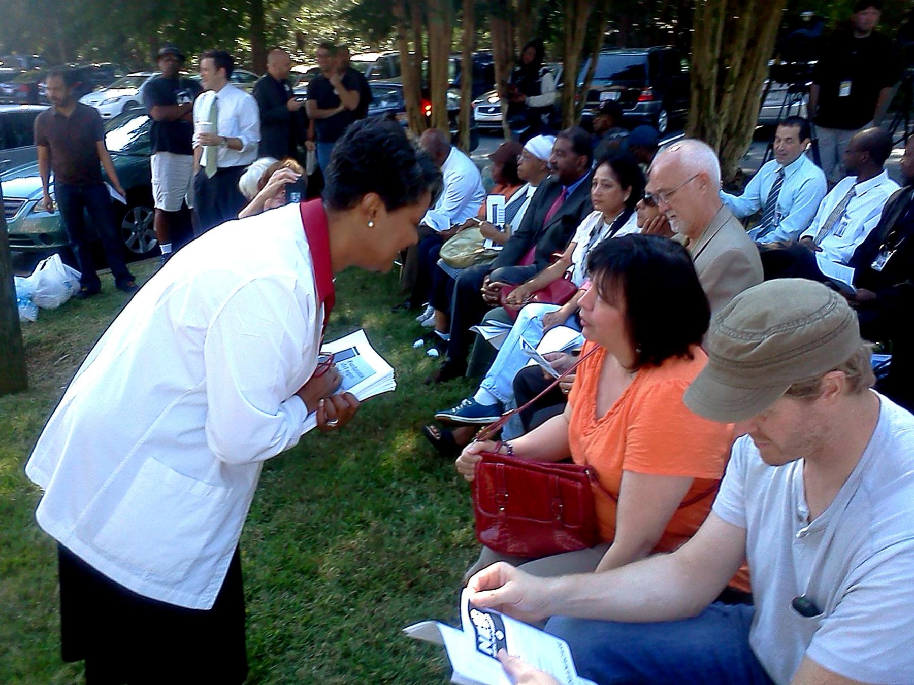 A speaker in a white coat addresses community members seated outdoors at a CHCC health forum.